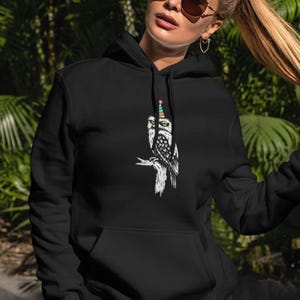 Op de afbeelding: Zwarte hoodie met een zwart-witte illustratie van een uil met een feesthoedje. De uil zit op een tak. De hoodie heeft een voorzak en een capuchon met trekkoord. De achtergrond toont wazig groen gebladerte.