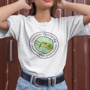 May include: White t-shirt with a circular graphic of a green and yellow snake. The graphic includes the text "Common Tree Snake" and "Dendrelaphis punctulatus". The shirt is made of a soft material.