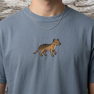 Op de afbeelding: Een blauwgrijs T-shirt met een geborduurd Thylacine, een uitgestorven buideldier. De Thylacine is bruin en zwart, met een gestreepte rug. Een zilveren ketting is zichtbaar. Het shirt is gemaakt van zacht materiaal.