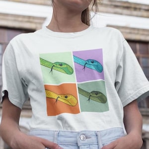 Puede incluir: Camiseta blanca con un diseño pop art de cuatro serpientes de colores. Cada serpiente es de un color diferente: verde, azul, amarillo y verde oliva, sobre un fondo pastel. Las serpientes tienen lenguas negras.