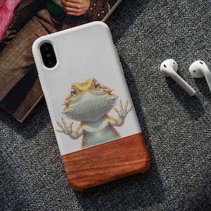 Può includere: Una custodia per smartphone bianca con un'illustrazione grafica di un drago barbuto giallo e arancione con gli artigli anteriori premuti contro la superficie. La parte inferiore della custodia è in legno marrone scuro con venature visibili.