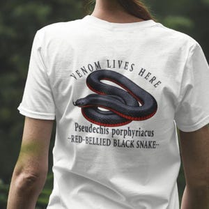 May include: White t-shirt with a graphic of a black snake with a red belly. The text "VENOM LIVES HERE" is above the snake, and "Pseudechis porphyriacus - RED-BELLIED BLACK SNAKE-" is below. A casual, everyday wear item.
