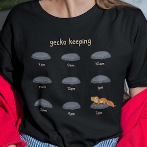 May include: A black t-shirt featuring the text gecko keeping above a grid of nine gray rocks labeled with times from 7am to 7pm. An orange gecko is illustrated resting on the rock labeled 7pm, while the other eight rocks remain empty.