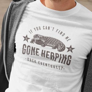 May include: White t-shirt with a lizard graphic and the text "IF YOU CAN'T FIND ME GONE HERPING - BACK EVENTUALLY". The design includes stars and a vintage aesthetic.