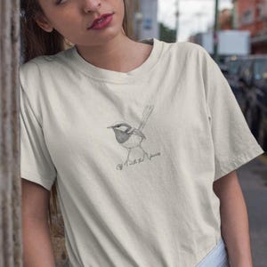Könnte beinhalten: Cremefarbenes T-Shirt mit einer Bleistiftskizze eines Vogels. Der Vogel sitzt auf einem Ast, darunter steht "Off with the fairies". Das T-Shirt hat kurze Ärmel und eine lockere Passform.
