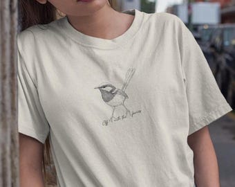 May include: Cream-colored t-shirt featuring a detailed pencil sketch of a bird. The bird is perched on a branch, with the words "Off with the fairies" written below it in a delicate script. The shirt has short sleeves.