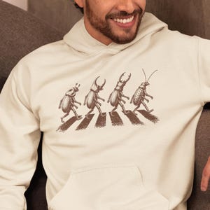 May include: Cream-coloured hoodie with a brown graphic of four beetles crossing a zebra crossing. The beetles are depicted walking, with the crossing stripes in a darker brown. The hoodie has a front pocket and a drawstring hood.