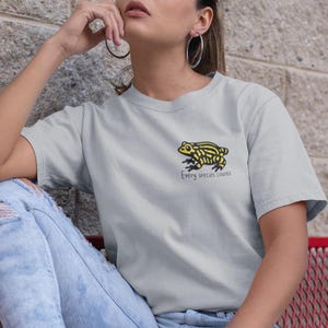 Peut inclure: Un t-shirt gris clair avec un motif de grenouille jaune et noir. Le texte "Every species counts" est imprimé en dessous de la grenouille. Le t-shirt est fait d'une matière douce.