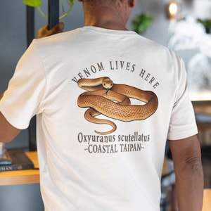 May include: Off-white t-shirt with a brown snake illustration and the text "VENOM LIVES HERE". The text "Oxyuranus scutellatus --COASTAL TAIPAN--" is below the snake graphic. The shirt is a light colour.