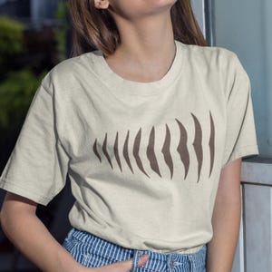Op de afbeelding: Beige T-shirt met korte mouwen en een bruin, abstract ontwerp van gebogen, verticale vormen. Het ontwerp bestaat uit een reeks langwerpige vormen. Het T-shirt is gemaakt van zacht materiaal.