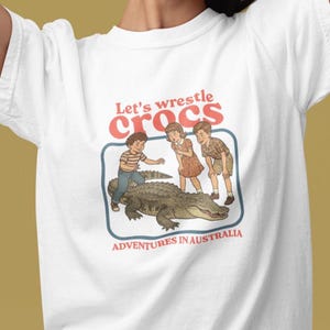 Könnte beinhalten: Weißes T-Shirt mit einer Grafik, die drei Kinder und ein Krokodil zeigt. Der Text "Let's wrestle crocs" steht über dem Bild, und "ADVENTURES IN AUSTRALIA" steht darunter.