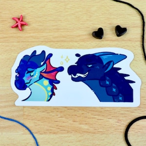 May include: A sticker depicting two cartoon dragons, one light blue with red and yellow details, the other dark blue. The sticker has a white border and is on a wooden surface with decorative items.