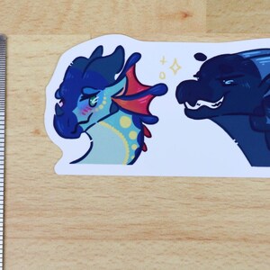 May include: A sticker featuring two cartoon dragon heads. One dragon is blue with pink cheeks and a red fin, the other is dark blue. The sticker is approximately 7.6 cm long.