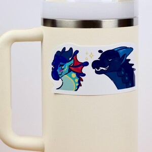 May include: A sticker depicting two cartoon dragons. One is blue and teal with red details, the other is dark blue. The sticker is attached to a cream-coloured travel mug with a silver rim and handle.
