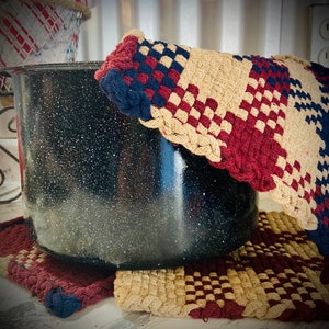May include: A black enamel pot with a speckled finish sits on a woven trivet with red, white, and blue checkered pattern. The trivet is made of cotton yarn.