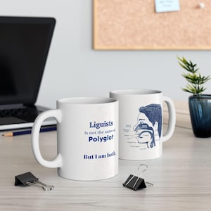 May include: Two white ceramic mugs with blue text and illustrations. The mug on the left says "Linguists is not the same as Polyglot But I am both." The mug on the right has a drawing of a person's head and neck with the different parts of the vocal tract labeled.