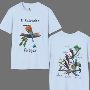 May include: Light blue t-shirt with a colorful bird illustration and the text "El Salvador Torogoz". The back of the shirt features a branch with colorful birds and the names of Central American countries.