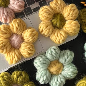May include: A collection of six crocheted flower embellishments in various colors, including yellow, pink, green, and light blue. The flowers are made with yarn and have a three-dimensional appearance.