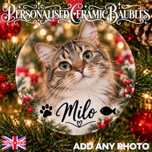 May include: A round ceramic ornament featuring a high-resolution photograph of a brown tabby cat. The text Milo is printed in black script, accompanied by a black paw print and fish icon. The ornament is displayed against a blurred Christmas tree.