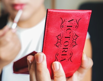 May include: A red leather compact mirror with the word "VICTORIA" in black lettering and a decorative design.