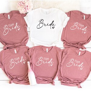Puede incluir: Cinco camisetas con el texto "Team Bride" en escritura blanca sobre un fondo rosa polvoriento. Una camiseta blanca con el texto "Bride" en escritura negra.