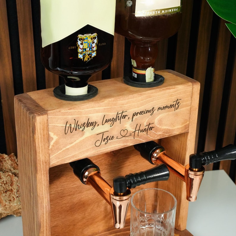 A wooden dispenser with two copper taps, one on each side, and a glass in front of it. The dispenser has a message engraved on it that says "Whiskey, laughter, precious moments Jose & Hunter".