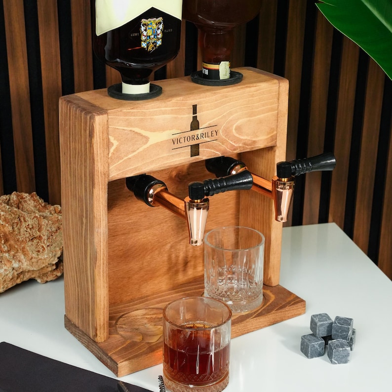 A wooden dispenser with two copper taps, likely for dispensing a beverage, is placed on a table with a glass of whiskey and some gray cubes nearby.