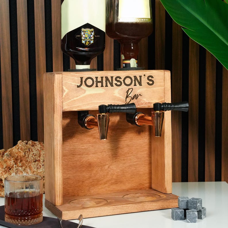A wooden bar tap with a personalized inscription, a bottle of whiskey, and a glass of whiskey on a wooden table.
