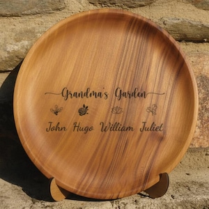 May include: A circular wooden serving platter, engraved with the words "Grandma's Garden" and the names John, Hugo, William, and Juliet. The tray displays a natural wood grain and is supported by a small wooden stand.