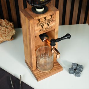 A wooden dispenser with a glass and copper-colored spout, featuring the name 'Daphne & Henry Wilson' on it. The dispenser is placed on a white surface, with a few small rocks nearby.