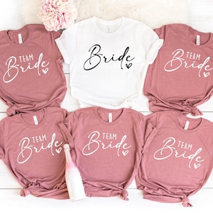 Pode incluir: Seis camisetas com o texto "Team Bride" em letras brancas sobre um fundo malva. Uma camiseta branca com o texto "Bride" em letras pretas.