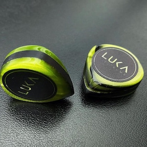 Puede incluir: Dos púas de guitarra verde y negro con la palabra "LUKA" impresa en oro en cada púa.