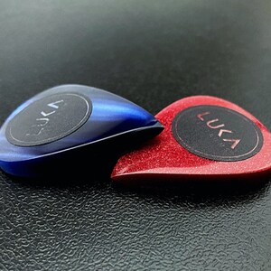 May include: Two guitar picks with a black circle in the center. The pick on the left is blue with a swirl pattern. The pick on the right is red with a sparkly finish. Both picks have the word "LUKA" printed in white on the black circle.