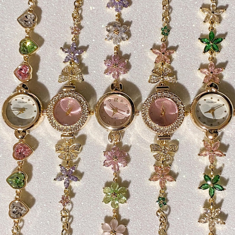 May include: Five gold-toned wristwatches with round faces and decorative floral and gemstone accents. The watch faces are pink and white, with the brand name "COLISEUM" visible on some. The bands feature heart, flower, and butterfly charms.