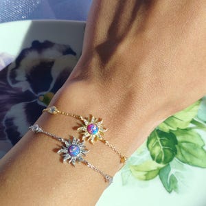 Puede incluir: Dos delicados brazaletes con dijes de sol de ópalo. El brazalete superior es de oro con un ópalo rosa, el brazalete inferior es de plata con un ópalo azul. Ambos brazaletes tienen pequeñas piedras transparentes alrededor de los dijes de sol.