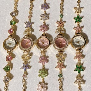 May include: Five gold-toned wristwatches with floral and gemstone accents. Each watch has a round face with a different colored dial. The watch bands feature chains, flowers, and gemstones in various colors, including pink, green, and purple.