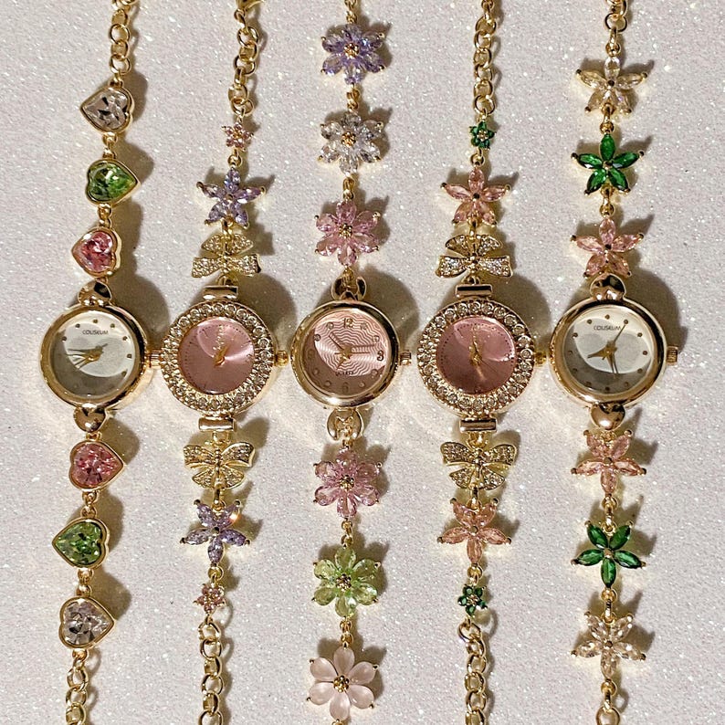 May include: Five gold-toned wristwatches with floral and gemstone accents. Each watch has a round face with a light-colored dial and delicate hands. The watch bands feature chains and are adorned with colorful flowers and heart-shaped gems.