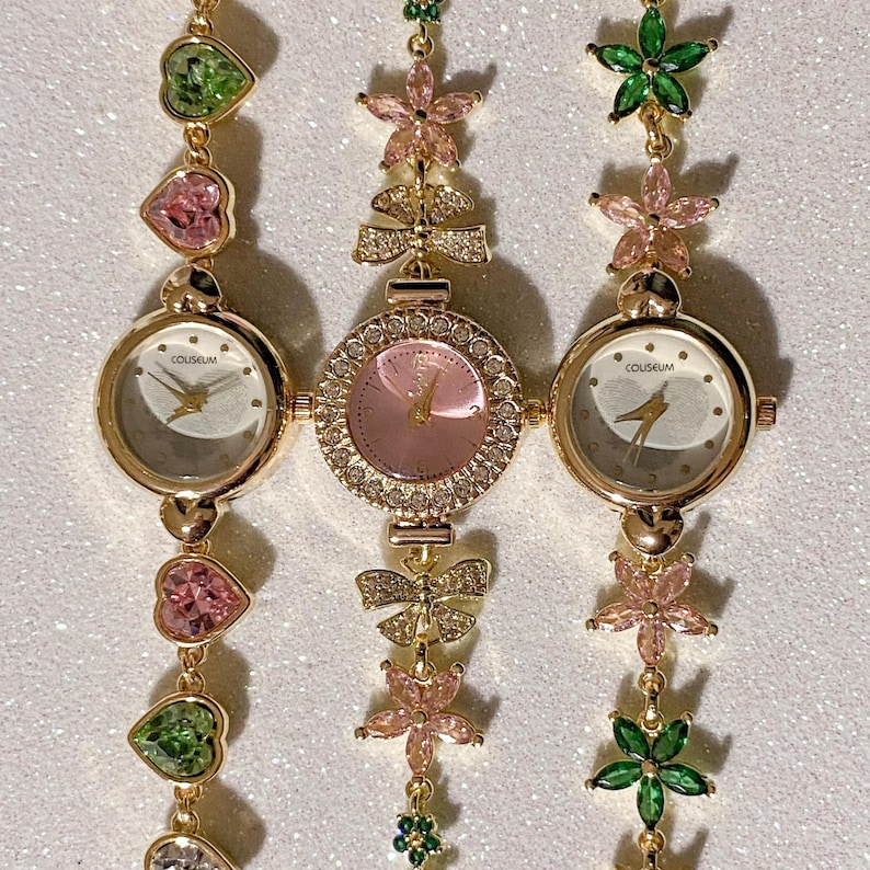 May include: Three gold-toned watches with white and pink faces, connected by decorative chains. The chains feature heart-shaped and flower-shaped gemstones in pink and green. The watch faces display the word "COLISEUM".
