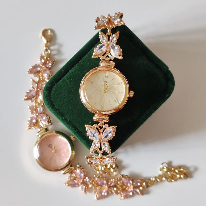 Puede incluir: Un reloj dorado con esfera color crema y una correa en forma de mariposa. La esfera del reloj tiene un dise&ntilde;o floral. Tambi&eacute;n se ve una pulsera a juego con detalles de piedras preciosas rosas. El reloj se exhibe sobre una superficie de terciopelo verde.