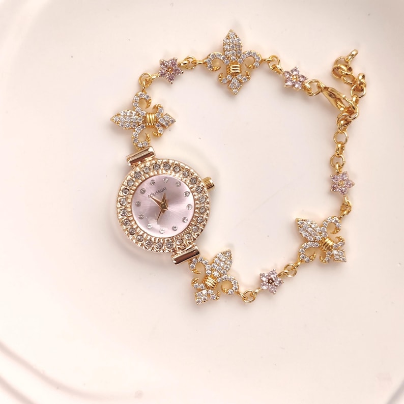 May include: A gold-tone watch with a white face and a crystal-encrusted bezel. The watch is attached to a gold chain bracelet with crystal-encrusted fleur-de-lis charms.