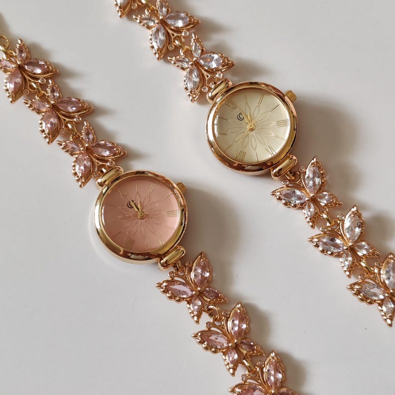Puede incluir: Dos relojes de pulsera dorados con dise&ntilde;os florales en las esferas y correas. Las correas est&aacute;n adornadas con detalles de mariposas de cristal rosa y transparente. Un reloj tiene una esfera rosa, el otro una esfera crema.