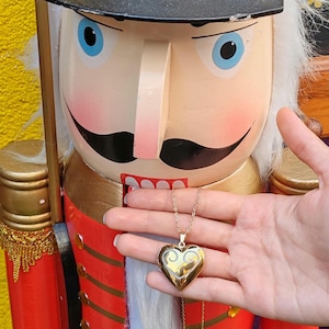 May include: A gold heart-shaped locket necklace with a decorative swirl design. The locket is suspended from a delicate gold chain. The background features a red and gold nutcracker.