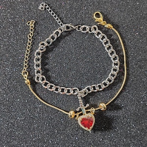 Op de afbeelding: Een zilveren enkelketting met een gouden enkelketting eraan bevestigd. De gouden ketting heeft een rode hartbedel met een zilveren zwaard erdoorheen.