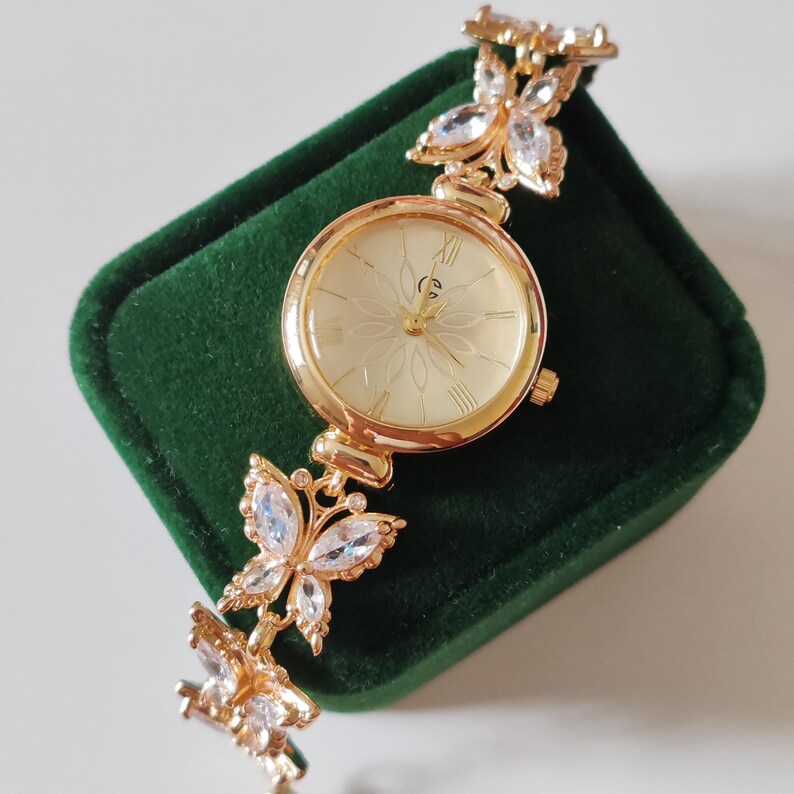 Puede incluir: Reloj de pulsera dorado con esfera crema y n&uacute;meros romanos. La correa del reloj presenta detalles en forma de mariposa adornados con cristales transparentes. El reloj se exhibe sobre una superficie de terciopelo verde oscuro.