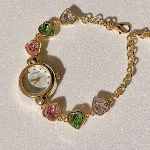 May include: Gold-toned bracelet watch with a round face and the word "COLISEUM" printed on the watch face. The bracelet features heart-shaped gemstones in pink, green, and clear colors.
