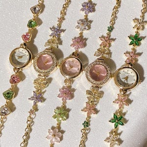 May include: Five gold-toned wristwatches with delicate floral and gemstone accents. Each watch features a round face with a different colored dial, including pink and white. The bands are adorned with colorful gemstones and flower charms.