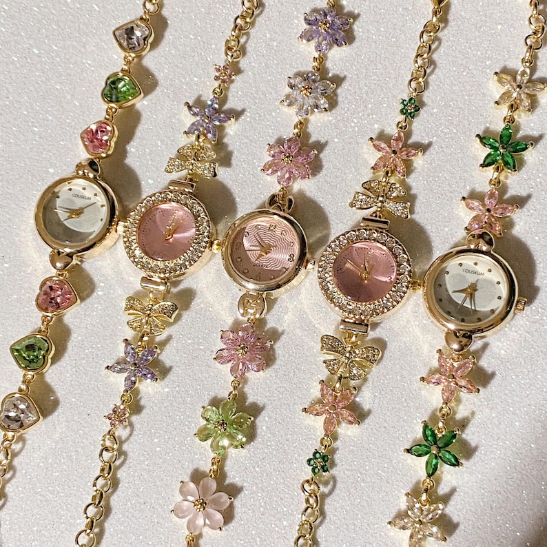 May include: Five gold-toned wristwatches with delicate floral and gemstone accents. Each watch features a round face with a different colored dial, including pink and white. The bands are adorned with colorful gemstones and flower charms.