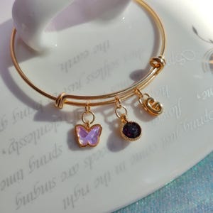 May include: A gold bangle bracelet with three charms: a purple butterfly, a dark purple gem, and a gold letter G.