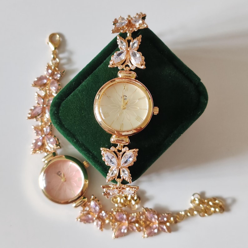 Puede incluir: Un reloj dorado con esfera crema y n&uacute;meros romanos, adornado con adornos en forma de mariposa. Tambi&eacute;n hay una pulsera a juego con detalles florales rosas. El reloj se exhibe sobre una superficie de terciopelo verde.