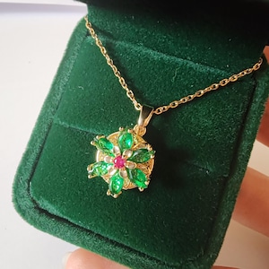 May include: A gold-colored necklace with a pendant shaped like a flower. The flower has green and red gemstones. The necklace is displayed in a green velvet jewelry box.